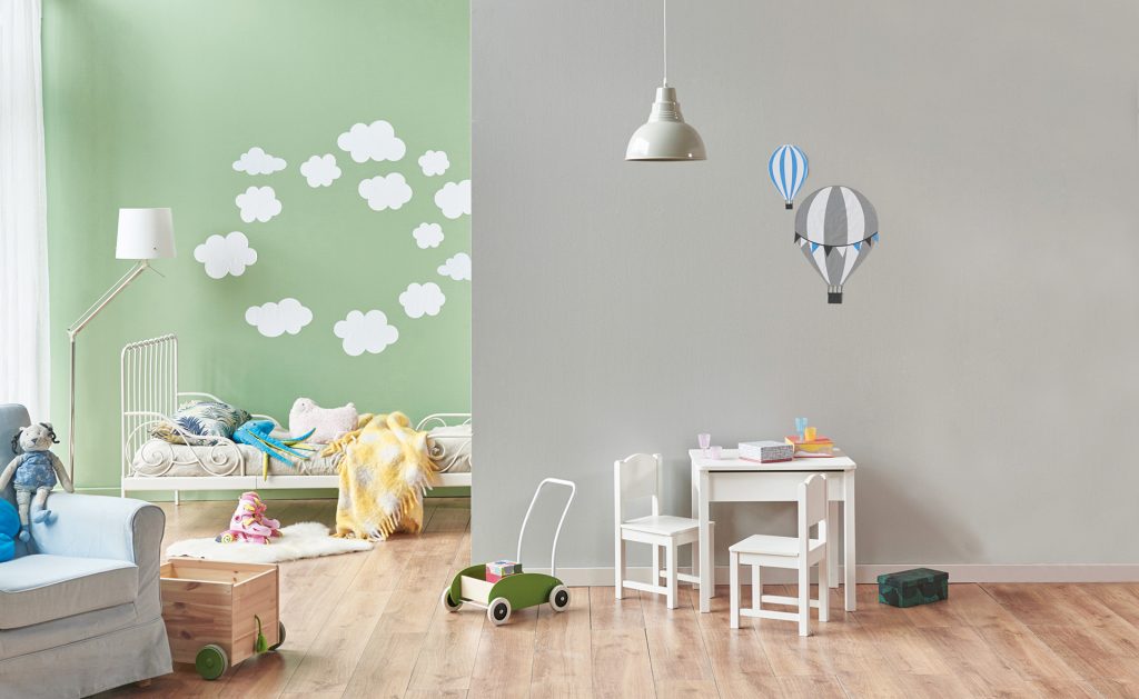 modernes Kinderzimmer mit einer gr&uuml;nen Wand mit wei&szlig;en Wolken und einer braunen Wand mit zwei aufgeklebten Hei&szlig;luftballons. Hinten steht ein wei&szlig;es Bett. Es gibt einen Sessel und eine kleinen wei&szlig;en Tisch mit 2 kleine wei&szlig;en St&uuml;hlen.